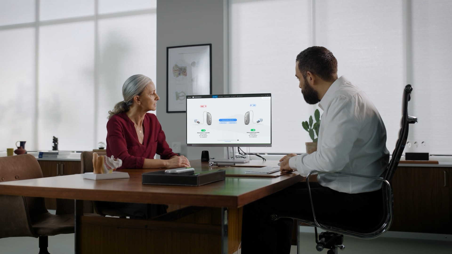 A female patient and a male hearing care professional sitting at a desk, reviewing hearing aid fitting software displayed on a computer monitor during a clinical consultation.