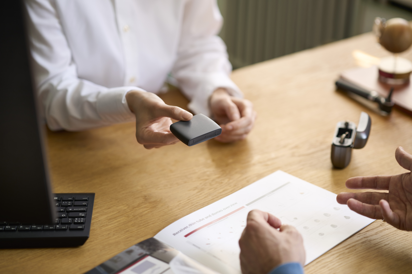 Two people at a table, one handing a hearing aid accessory to the other.