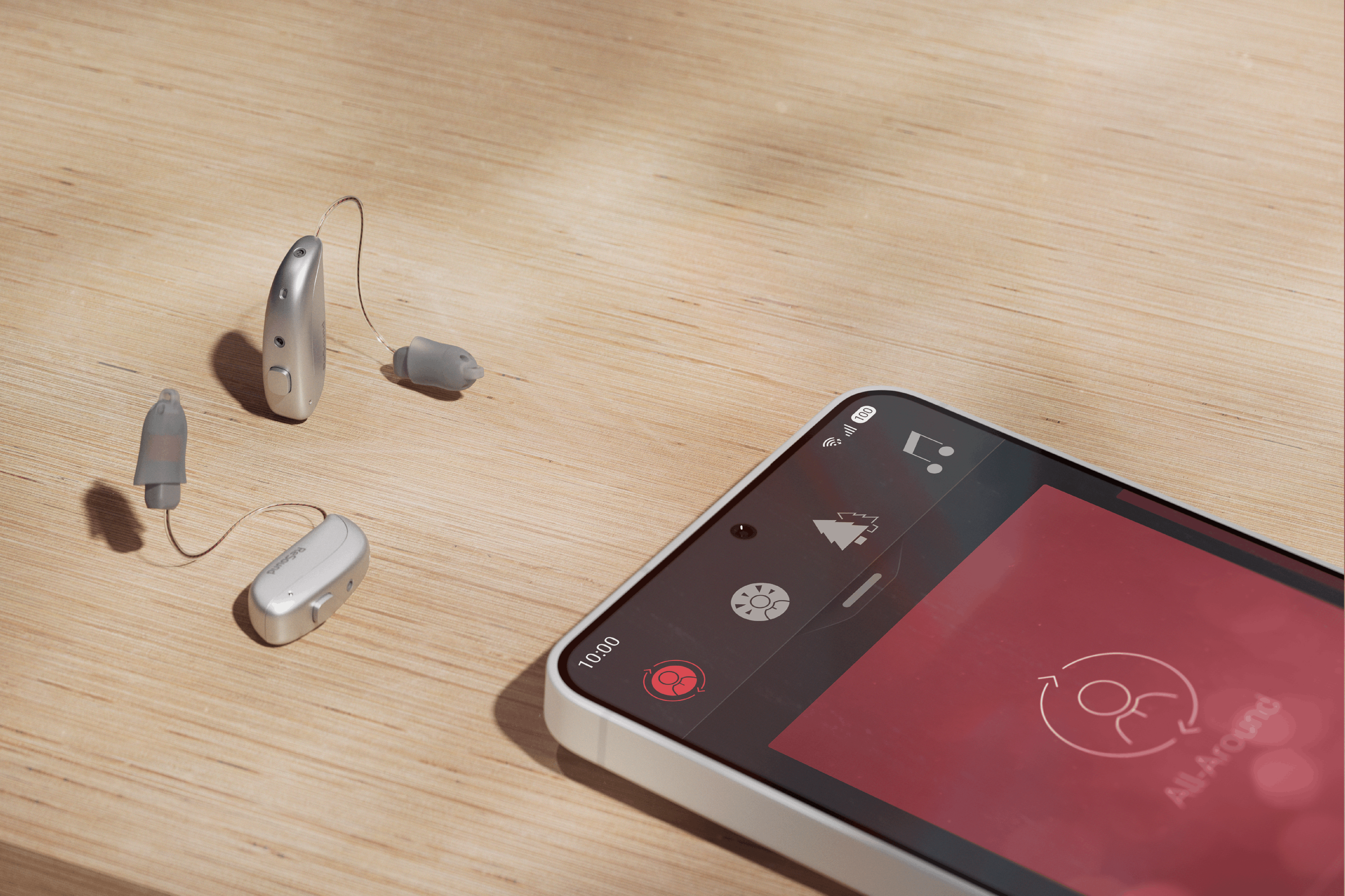 A pair of silver ReSound Sensia hearing aids next to a smartphone displaying the Smart 3D app on a light wood surface